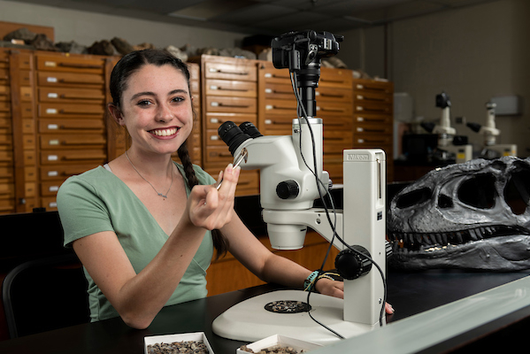 Emma Puetz sits in front of a microscope with dinosaur fossils and smiles at the camera